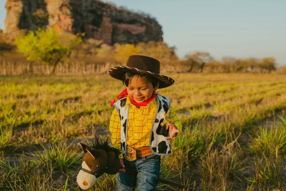 A young boy in a cowboy costume joyfully plays outdoors with a toy horse.