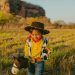 A young boy in a cowboy costume joyfully plays outdoors with a toy horse.
